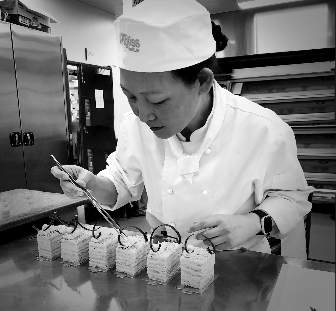 A black and white photograph of Yawen finishing off 6 small French Pastries in a commercial kitchen